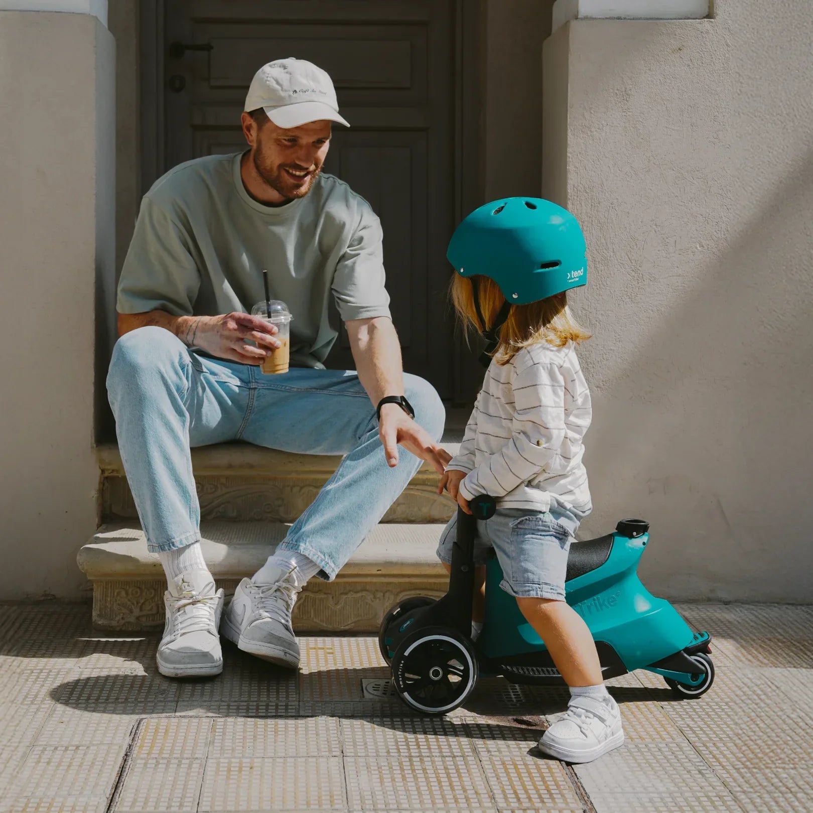 papá sentado en el pórtico de una casa junto a su hija pequeña con casco verde, montada en scooter mini ride-on de smartrike color verde 