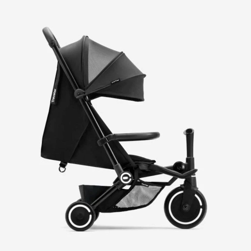Vista lateral del coche triciclo SmarTrike Traveler negro desplegado en modo paseo, mostrando toldo, respaldo reclinable, barra delantera, canasta de almacenamiento inferior con malla y tres ruedas con aros blancos. Fondo gris claro.