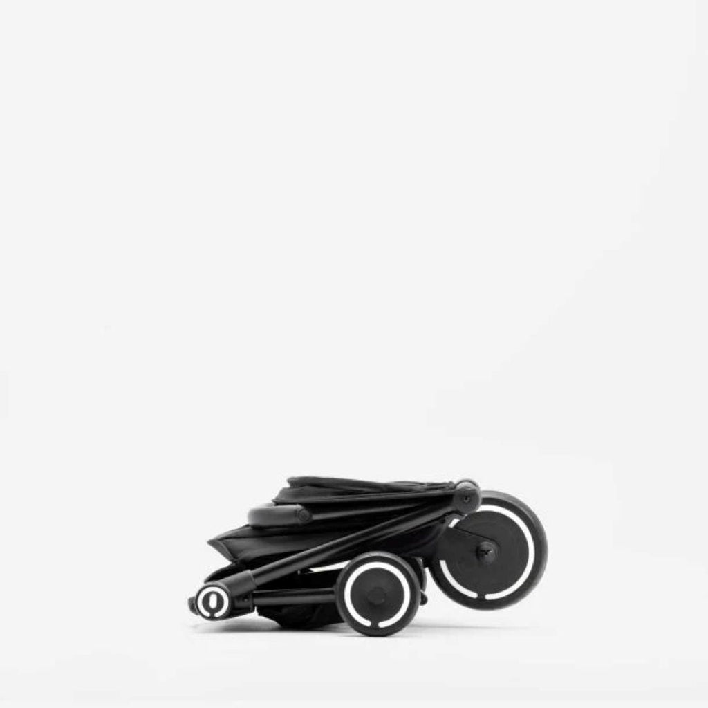 Vista lateral del coche triciclo SmarTrike Traveler negro completamente plegado en tamaño ultracompacto, con las tres ruedas visibles y aros blancos. Fondo blanco.