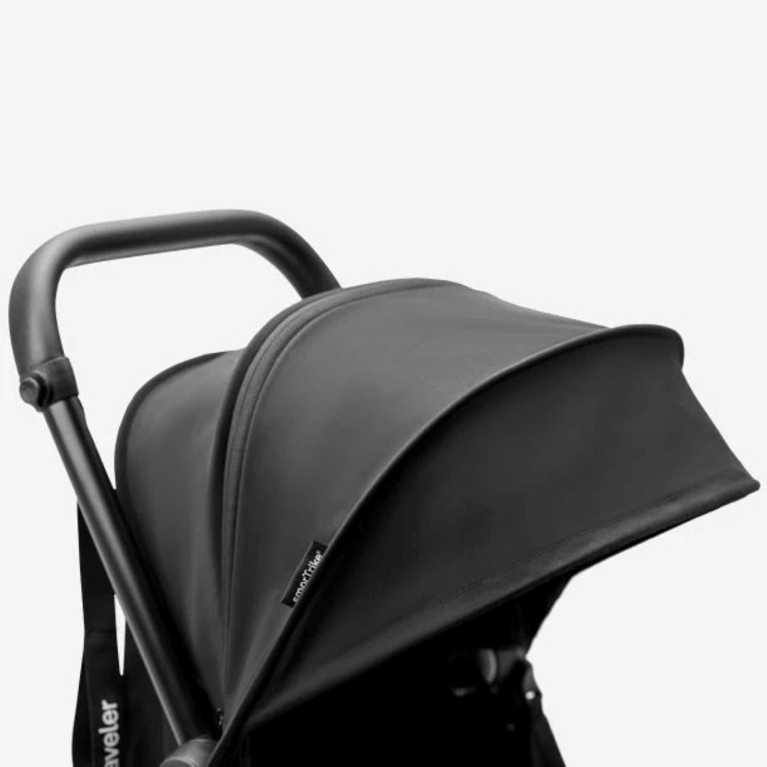 Detalle en primer plano del toldo solar negro de tela premium del coche triciclo SmarTrike Traveler, con manubrio curvo y etiqueta de la marca visible. Fondo blanco.