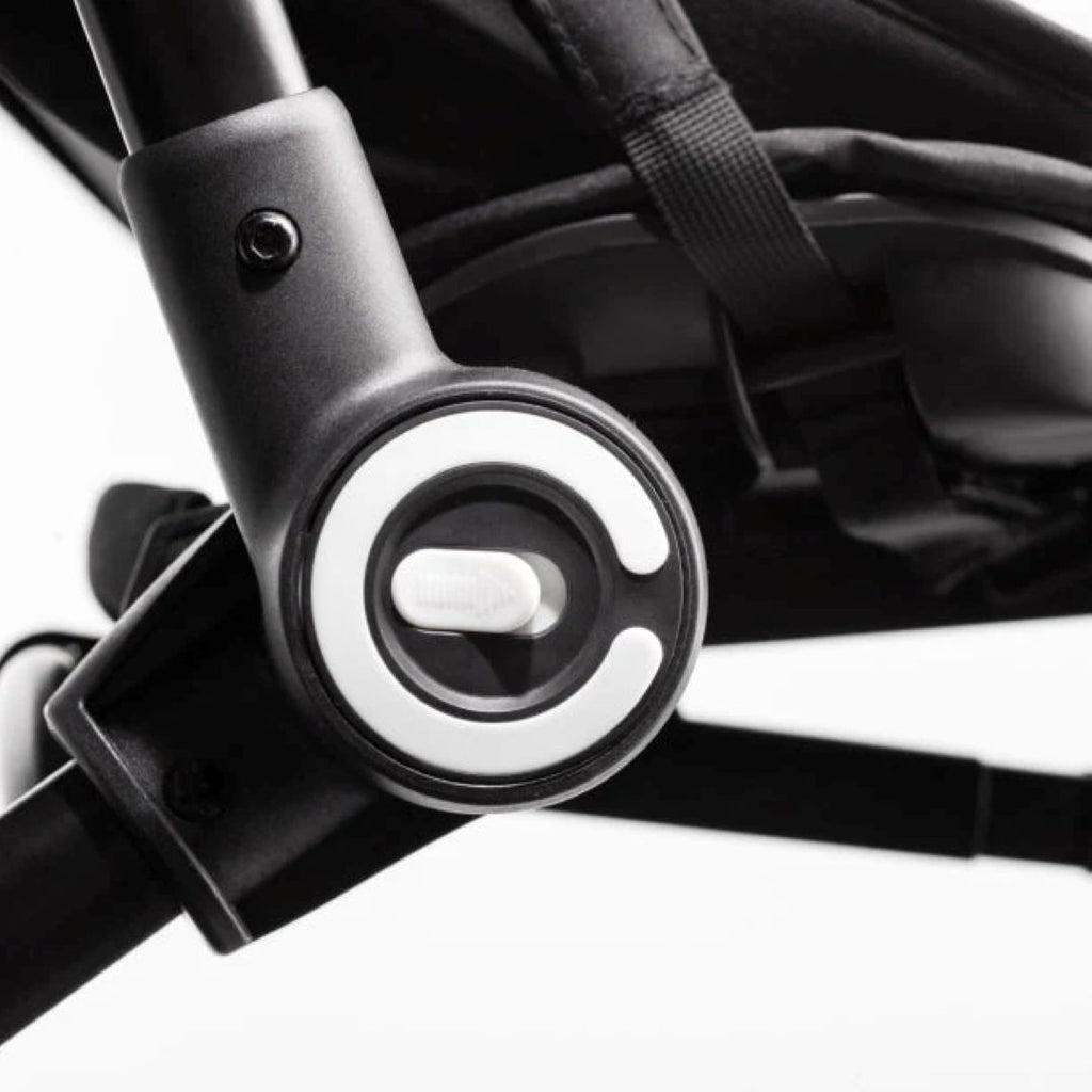 Detalle en primer plano del botón de plegado circular con logo SmarTrike del coche triciclo Traveler, sobre estructura negra. Fondo blanco.