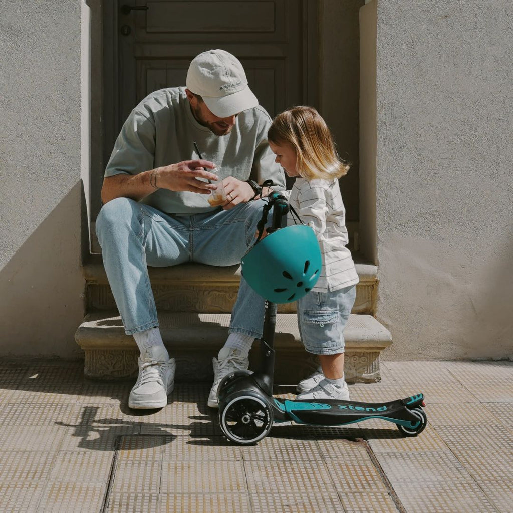 Papá sentado en escalones ayudando a su hija a ponerse el casco verde azulado, con el scooter SmarTrike Xtend Ride-On Petróleo junto a ellos. Contexto urbano soleado.