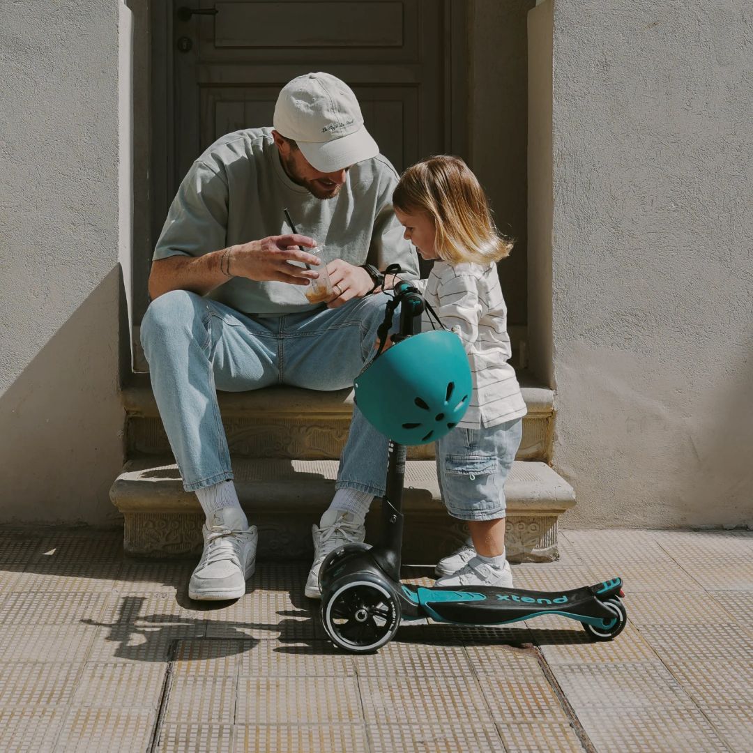 Papá sentado en escalones ayudando a su hija a ponerse el casco verde azulado, con el scooter SmarTrike Xtend Ride-On Petróleo junto a ellos. Contexto urbano soleado.