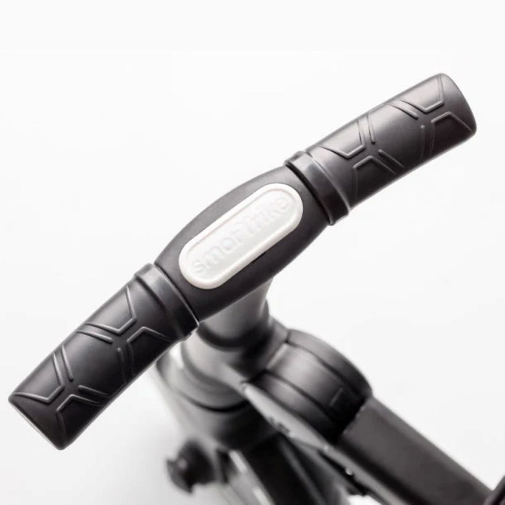 Detalle en primer plano del manubrio ergonómico negro con textura antideslizante del coche triciclo SmarTrike Traveler, con logo de la marca en relieve. Fondo blanco.