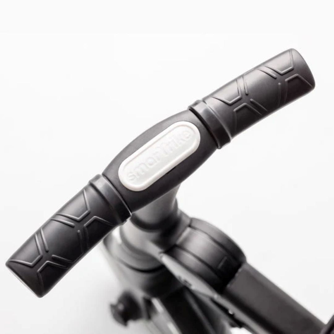 Detalle en primer plano del manubrio ergonómico negro con textura antideslizante del coche triciclo SmarTrike Traveler, con logo de la marca en relieve. Fondo blanco.