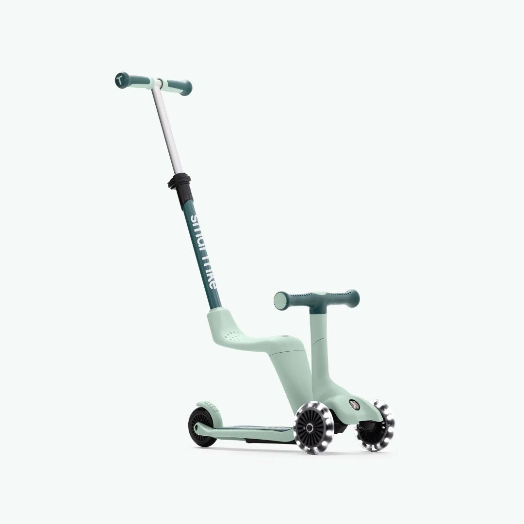 Vista frontal en ángulo del scooter evolutivo SmarTrike Xtend Mini Ride en color Verde Agua en modo correpasillos, con asiento integrado, manubrio para niño, barra para padres inclinada y tres ruedas con luz LED. Fondo verde muy claro.