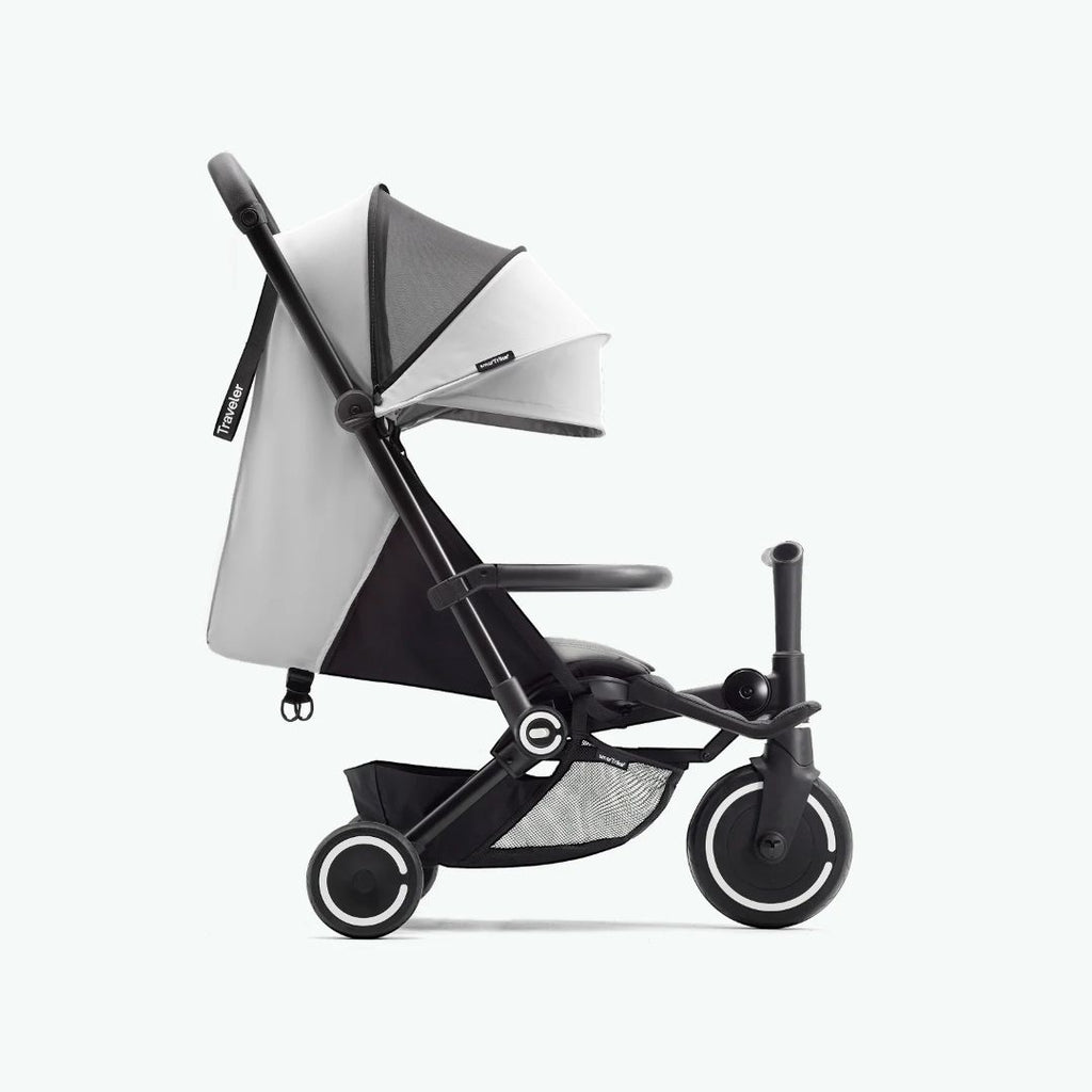 Vista lateral del coche triciclo SmarTrike Traveler gris claro desplegado en modo paseo, mostrando toldo, respaldo reclinable, barra delantera, canasta de almacenamiento inferior con malla y tres ruedas con aros blancos. Fondo gris claro.