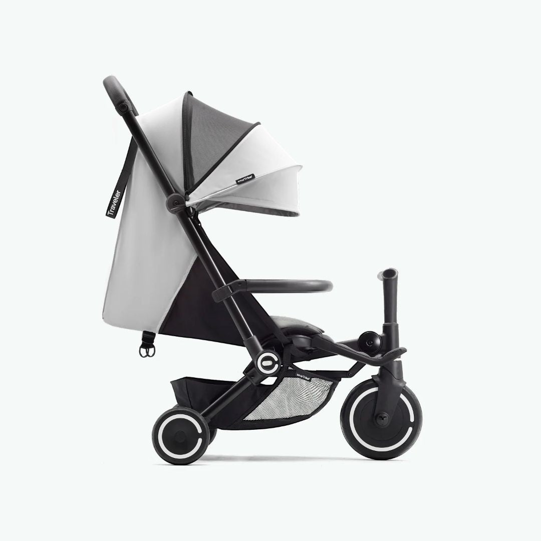Vista lateral del coche triciclo SmarTrike Traveler gris claro desplegado en modo paseo, mostrando toldo, respaldo reclinable, barra delantera, canasta de almacenamiento inferior con malla y tres ruedas con aros blancos. Fondo gris claro.