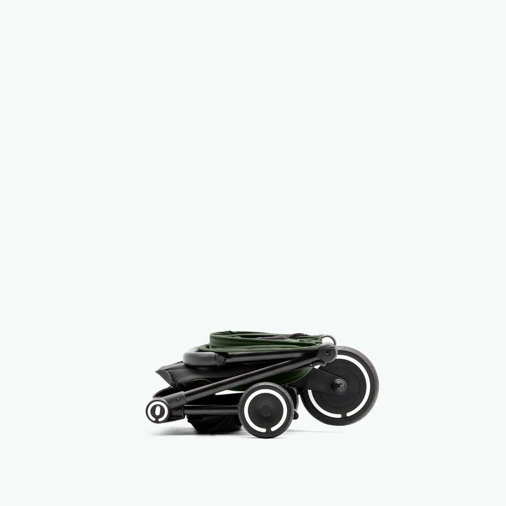 Vista lateral del coche triciclo SmarTrike Traveler verde completamente plegado en tamaño ultracompacto, con las tres ruedas visibles y aros blancos. Fondo verde muy claro.