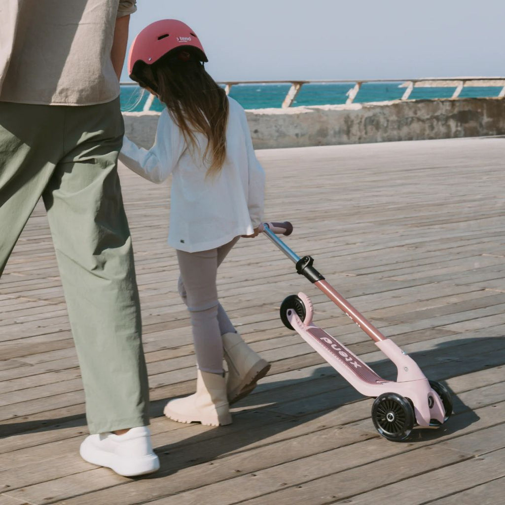 Niña con casco rosa caminando junto a un adulto mientras arrastra el scooter SmarTrike Xtend Mini+ en color Palo Rosa por un muelle de madera frente al mar.