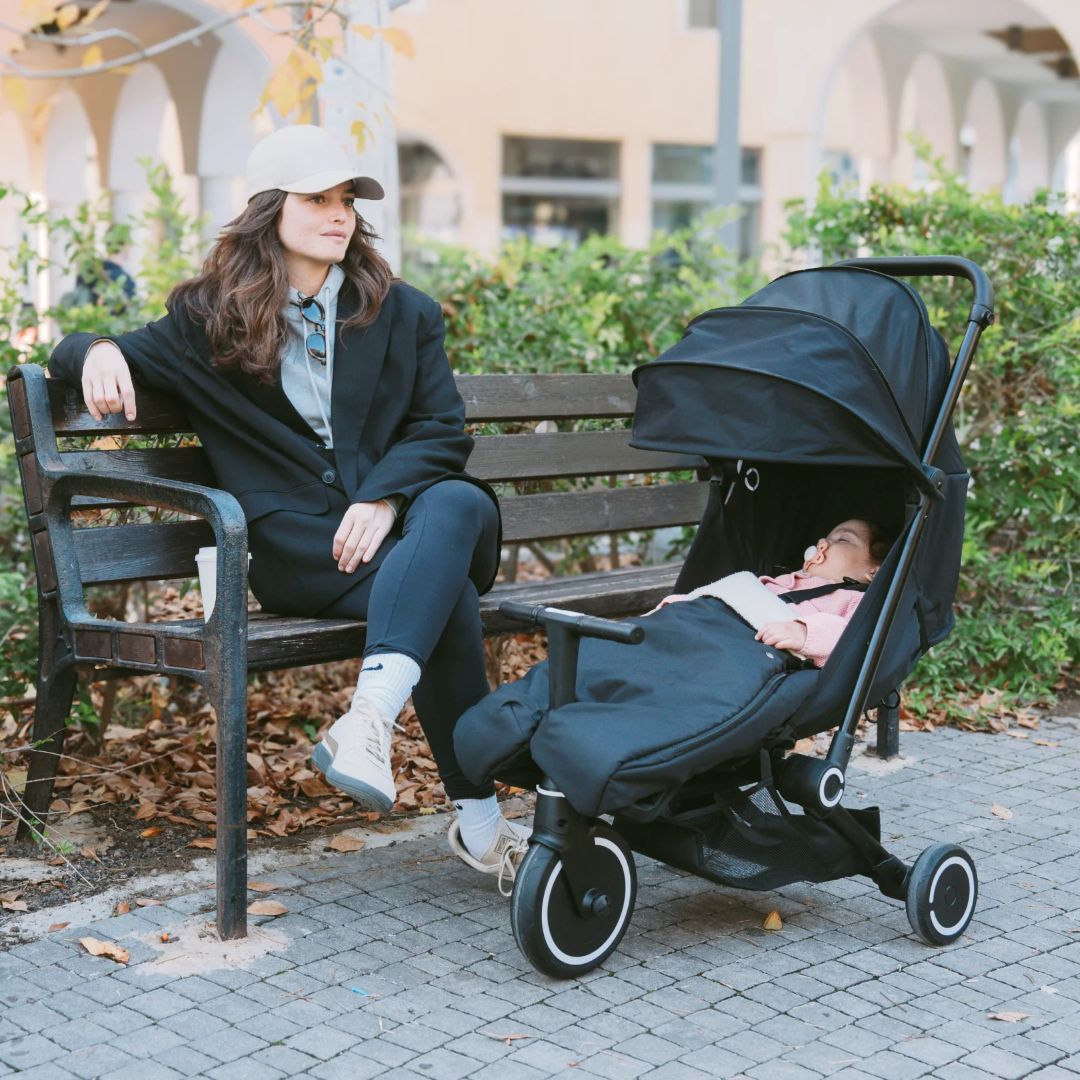 Mamá sentada relajada en una banca de parque mientras su bebé duerme reclinado en el coche triciclo SmarTrike Traveler negro con toldo extendido. Contexto urbano otoñal con hojas en el suelo.