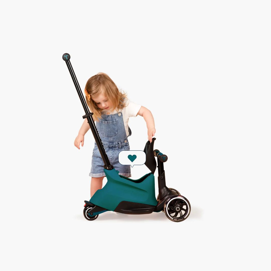 Niña pequeña de pie inclinándose curiosa sobre el scooter SmarTrike Xtend Ride-On en color verde azulado en modo ride-on, con ícono de corazón superpuesto. Fondo blanco.