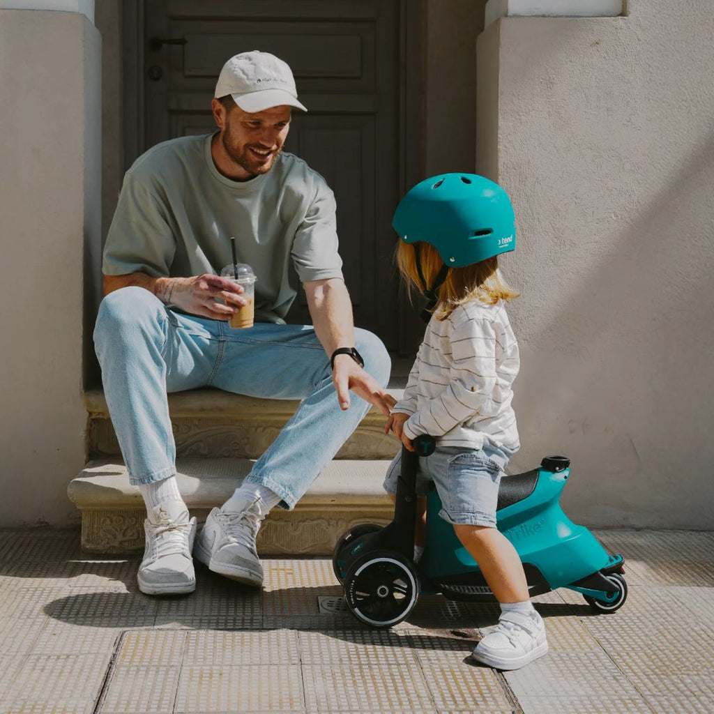 Papá sentado en unos escalones con café en mano sonriendo a su hija con casco verde azulado que monta el scooter SmarTrike Xtend Ride-On en modo ride-on, en una vereda soleada.