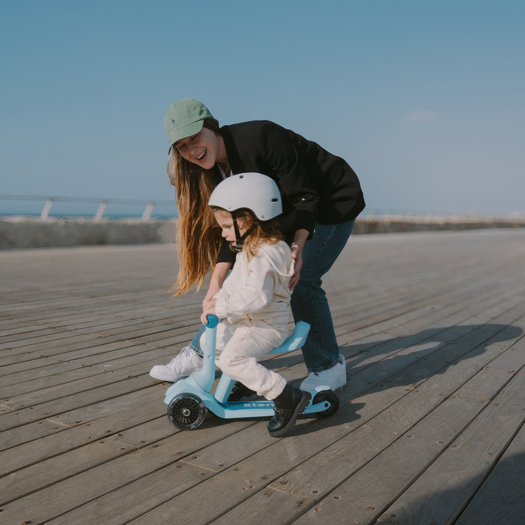 Mamá sonriente inclinada ayudando a su bebé con casco blanco a usar el scooter SmarTrike Xtend Mini Ride Celeste en modo correpasillos, sobre muelle de madera con cielo azul de fondo.