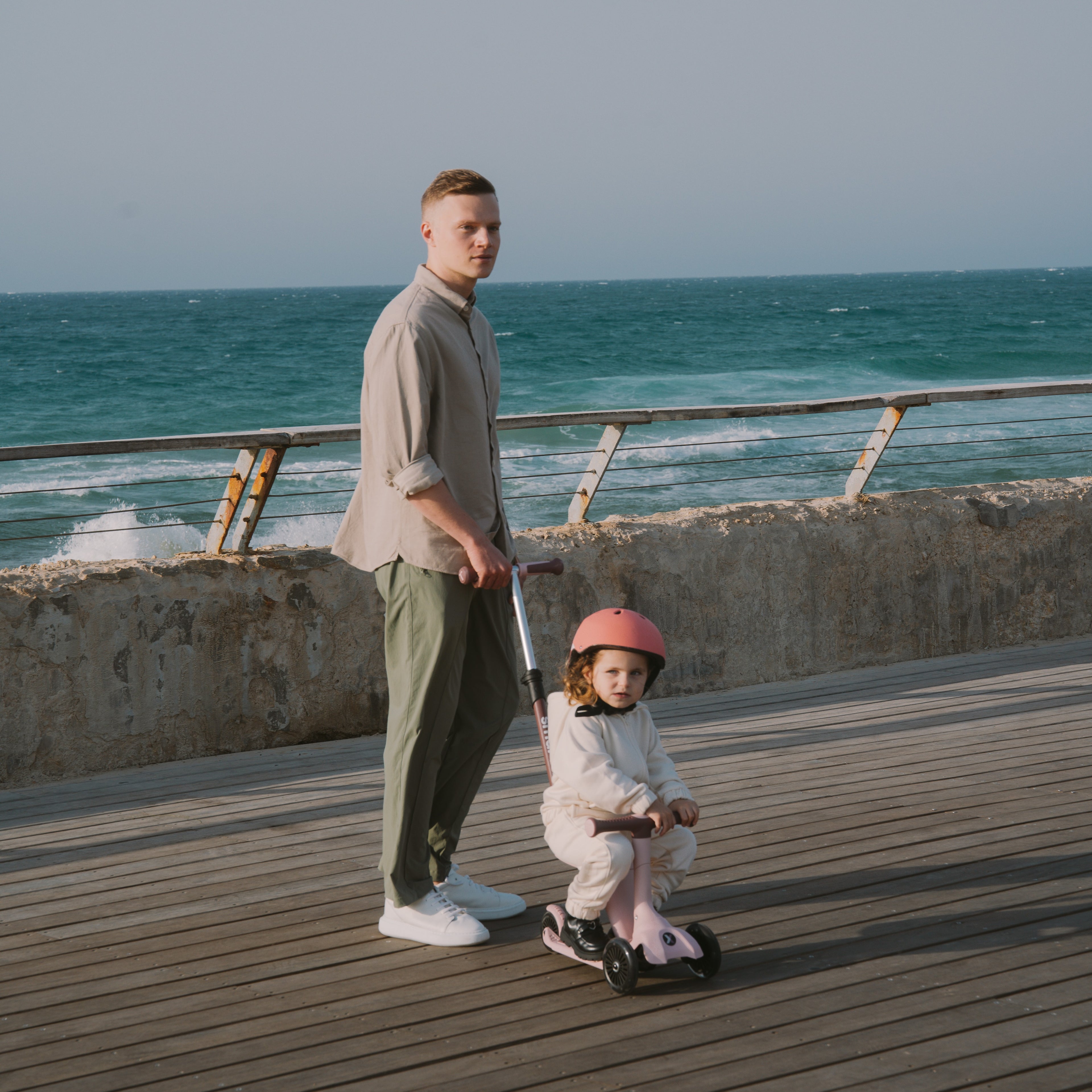 Papá de pie sosteniendo el manubrio del scooter SmarTrike Xtend Mini Ride Palo Rosa en modo control parental, mientras su hija pequeña con casco rosa va sentada en modo correpasillos, en un muelle frente al mar.