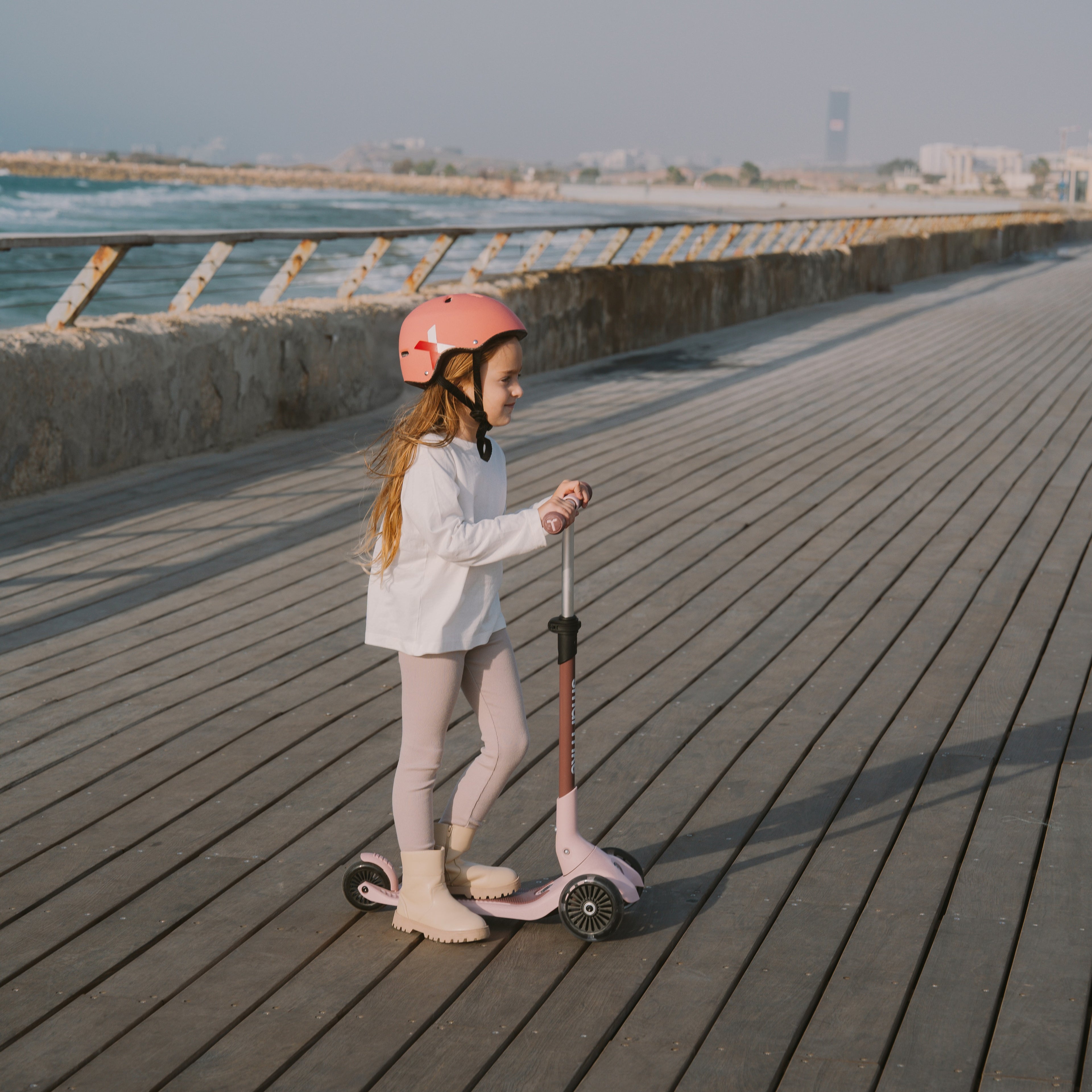 Niña de pie con casco rosa manejando de forma autónoma el scooter SmarTrike Xtend Mini Ride Palo Rosa osa en modo scooter, sobre muelle de madera con vistas al mar al atardecer.