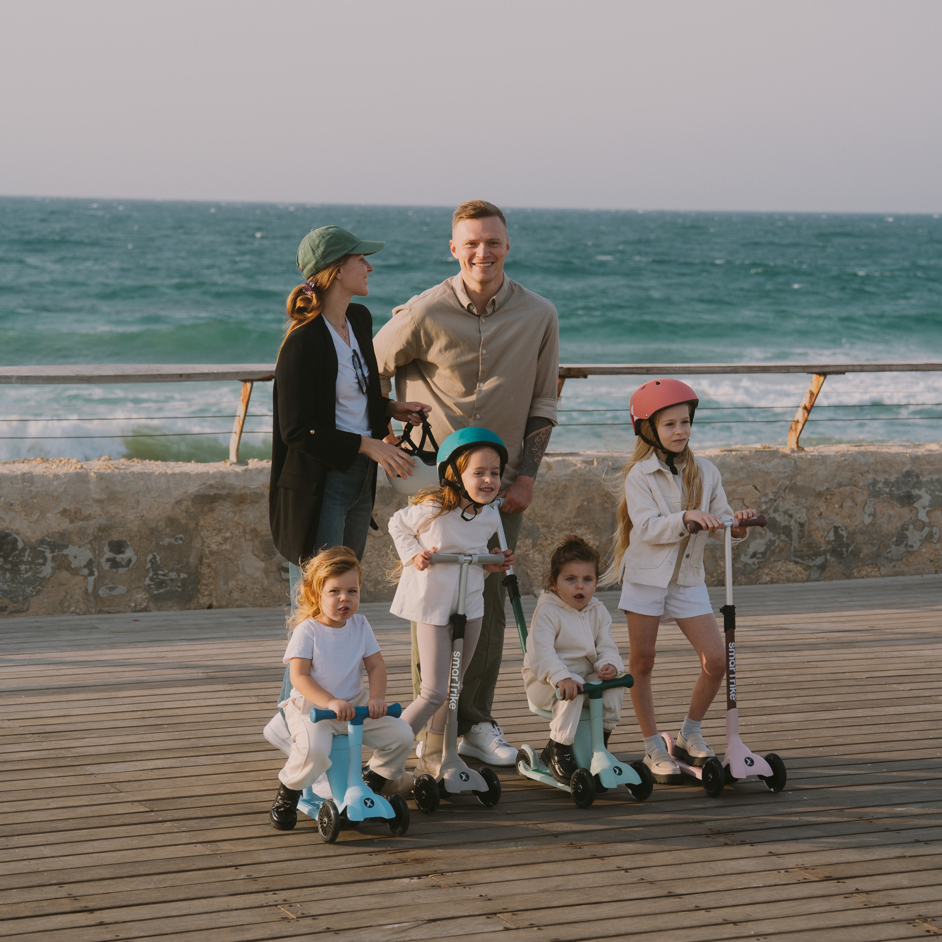 Familia con dos padres y cuatro niños posando en un muelle al atardecer frente al mar, con tres scooters SmarTrike Xtend Mini Ride en distintos colores y modos de uso.
