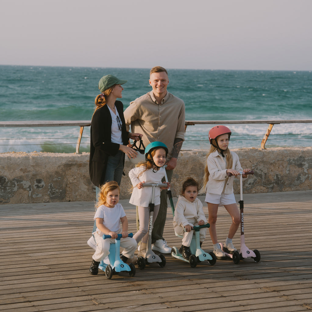Familia con dos padres y cuatro niños posando en un muelle al atardecer frente al mar, con tres scooters SmarTrike Xtend Mini Ride en distintos colores y modos de uso.