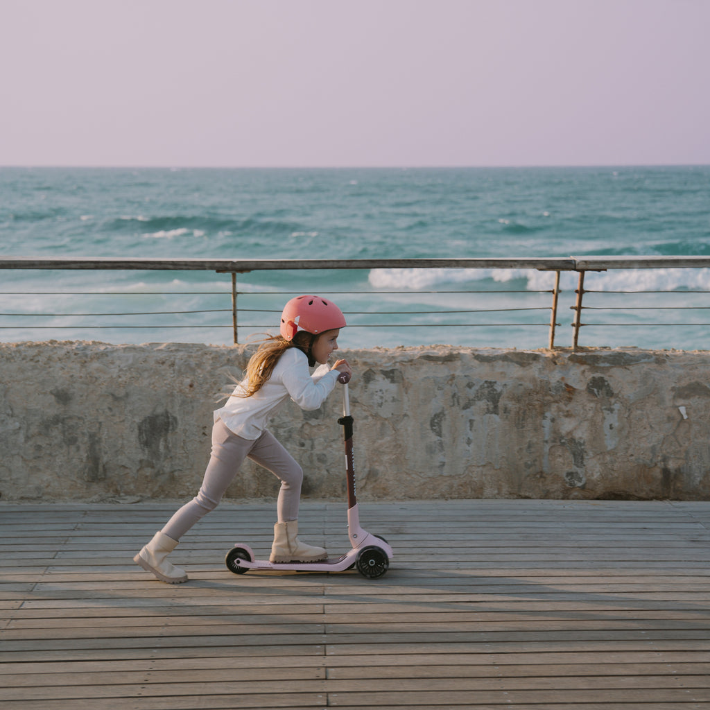 Niña con casco rosa impulsándose con energía en el scooter SmarTrike Xtend Mini+ Palo Rosa en modo scooter, sobre muelle de madera con el mar de fondo.