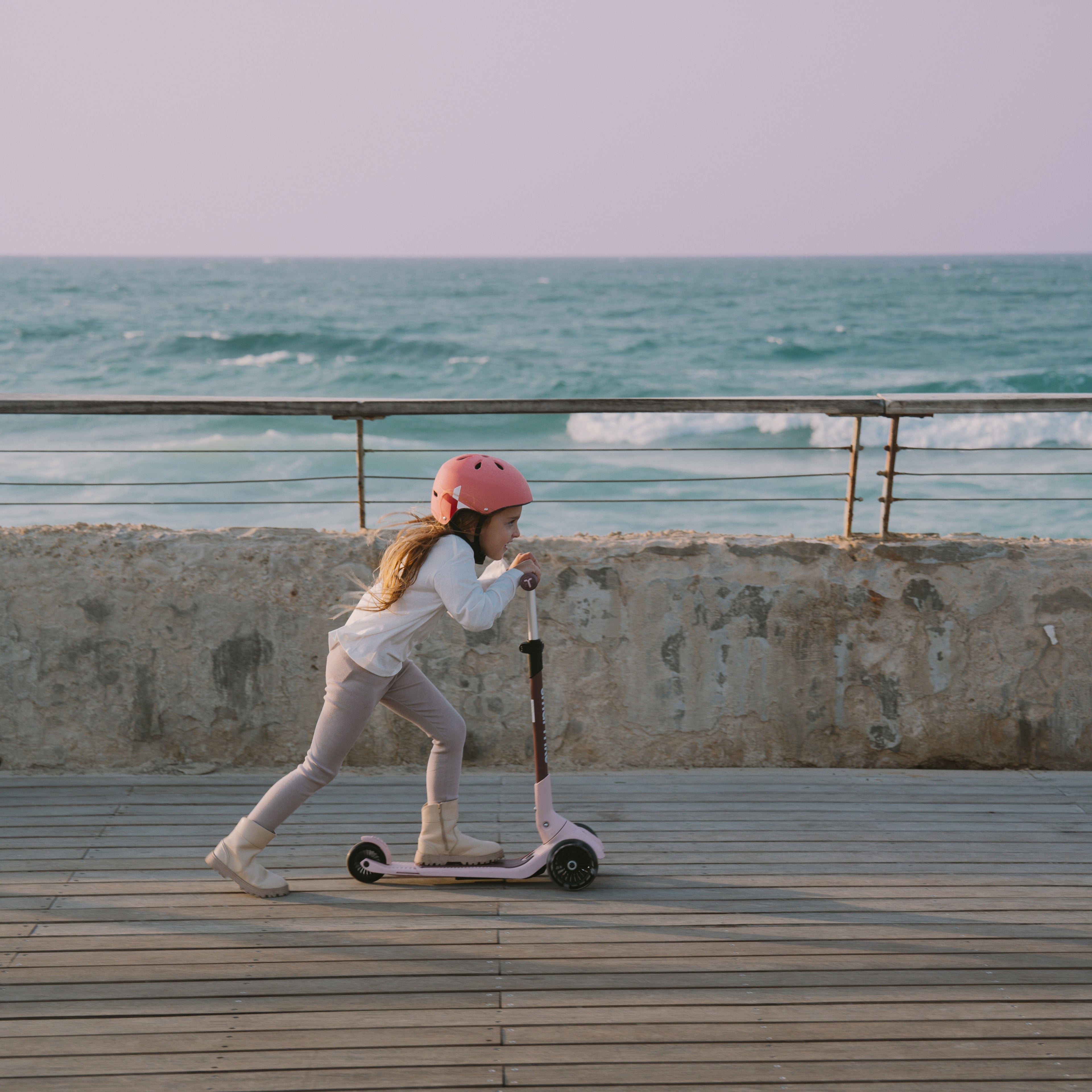 Niña con casco rosa impulsándose con energía en el scooter SmarTrike Xtend Mini+ Palo Rosa en modo scooter, sobre muelle de madera con el mar de fondo.