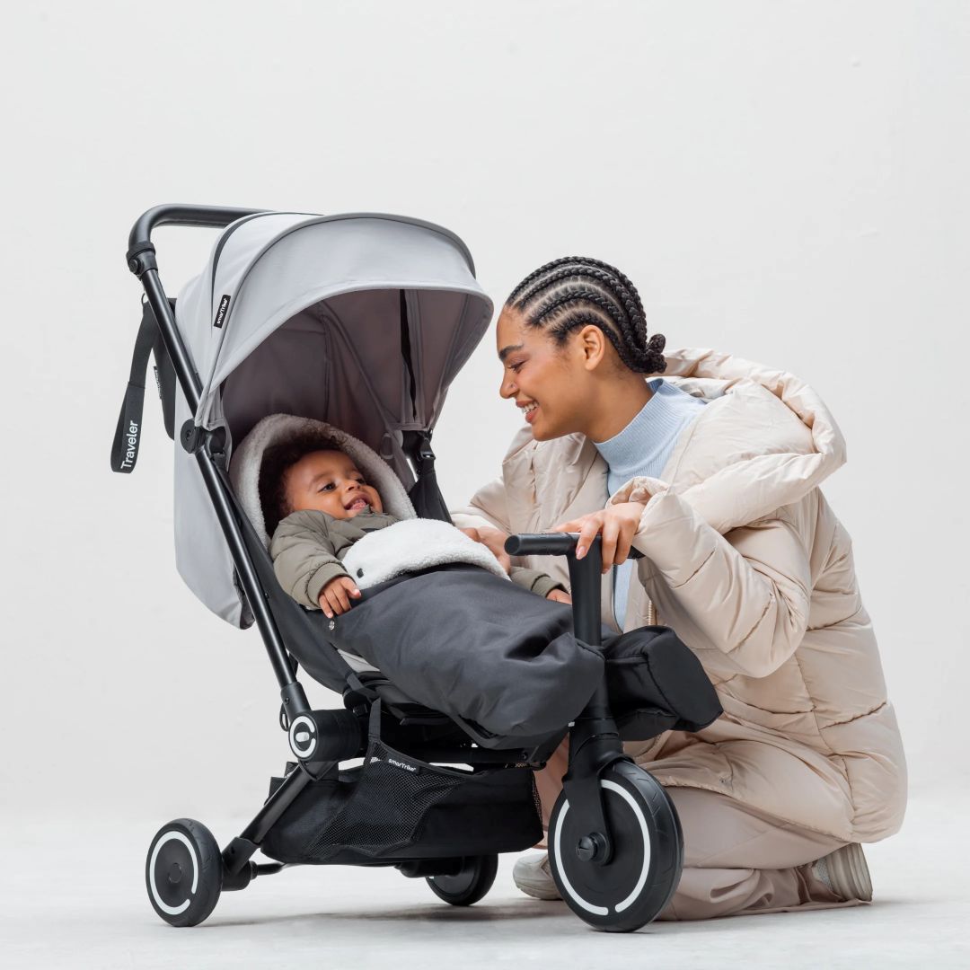 Bebé usando el coche triciclo Traveler de smarTrike con saco cubrepiés puesto, mamá sonriendo al lado