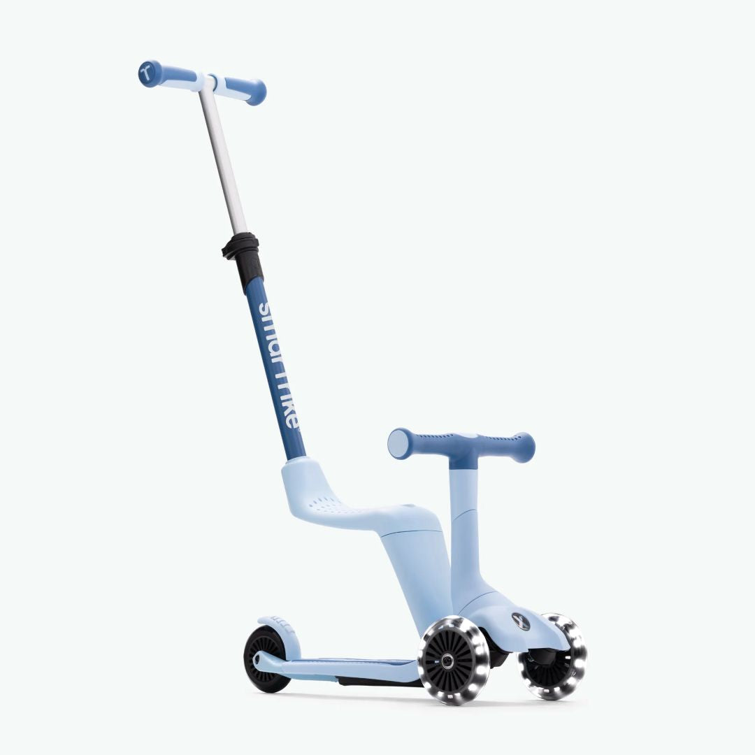 Vista frontal en ángulo del scooter evolutivo SmarTrike Xtend Mini Ride en color Celeste en modo correpasillos, con asiento integrado, manubrio para niño, barra para padres inclinada y tres ruedas con luz LED. Fondo celeste claro.