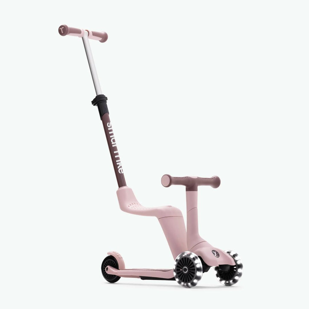 Vista frontal en ángulo del scooter evolutivo SmarTrike Xtend Mini Ride en color Palo Rosa en modo correpasillos, con asiento integrado, manubrio para niño, barra para padres inclinada y tres ruedas con luz LED. Fondo verde muy claro.