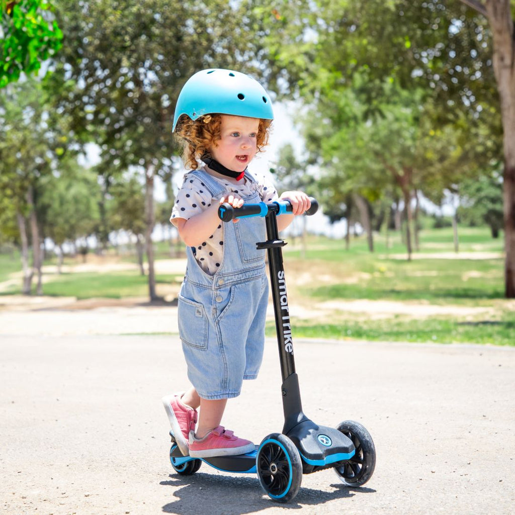 Bebé de aproximadamente 2 años con casco celeste de pie sobre el scooter evolutivo SmarTrike Xtend azul en etapa inicial, en un sendero de parque soleado con árboles de fondo.