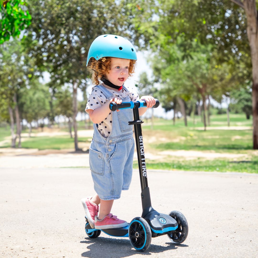 Bebé de aproximadamente 2 años con casco celeste de pie sobre el scooter evolutivo SmarTrike Xtend azul en etapa inicial, en un sendero de parque soleado con árboles de fondo.