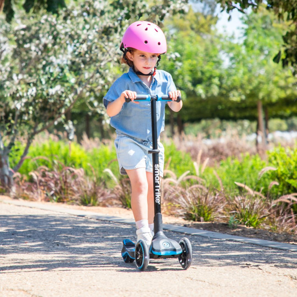 Niña de aproximadamente 5 años con casco rosado manejando el scooter evolutivo SmarTrike Xtend en color azul en un sendero de parque con árboles y vegetación de fondo.