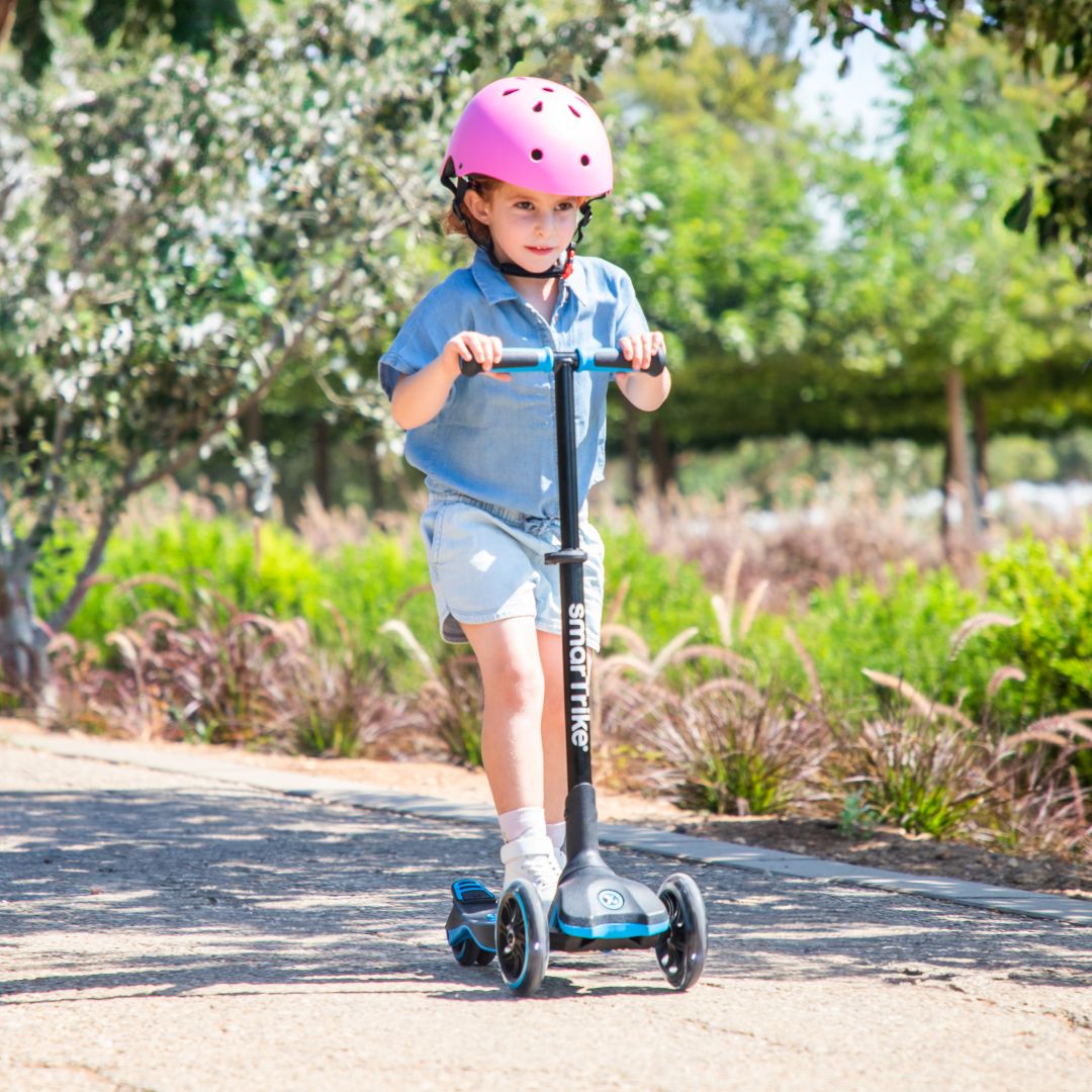 Niña de aproximadamente 5 años con casco rosado manejando el scooter evolutivo SmarTrike Xtend en color azul en un sendero de parque con árboles y vegetación de fondo.
