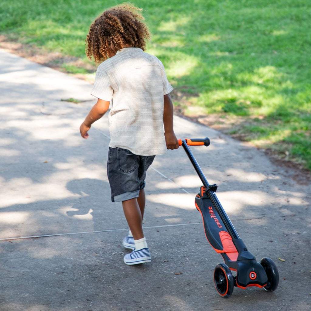  Niño pequeño de espaldas caminando y jalando el scooter SmarTrike Xtend naranja plegado por un sendero de parque soleado con césped de fondo.