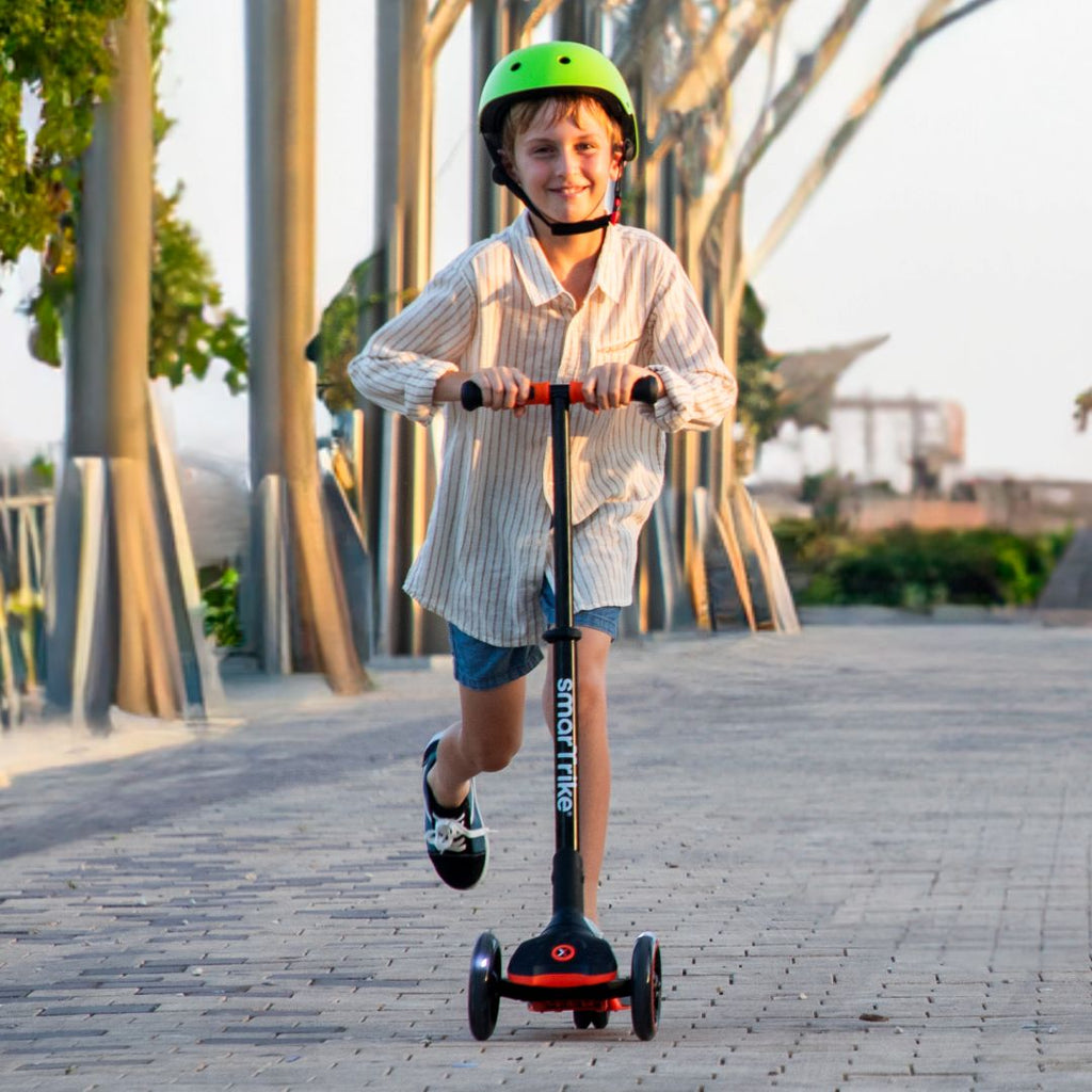 Niño de aproximadamente 9 años con casco verde sonriendo mientras maneja el scooter SmarTrike Xtend naranja en un paseo urbano al atardecer con arquitectura moderna de fondo.