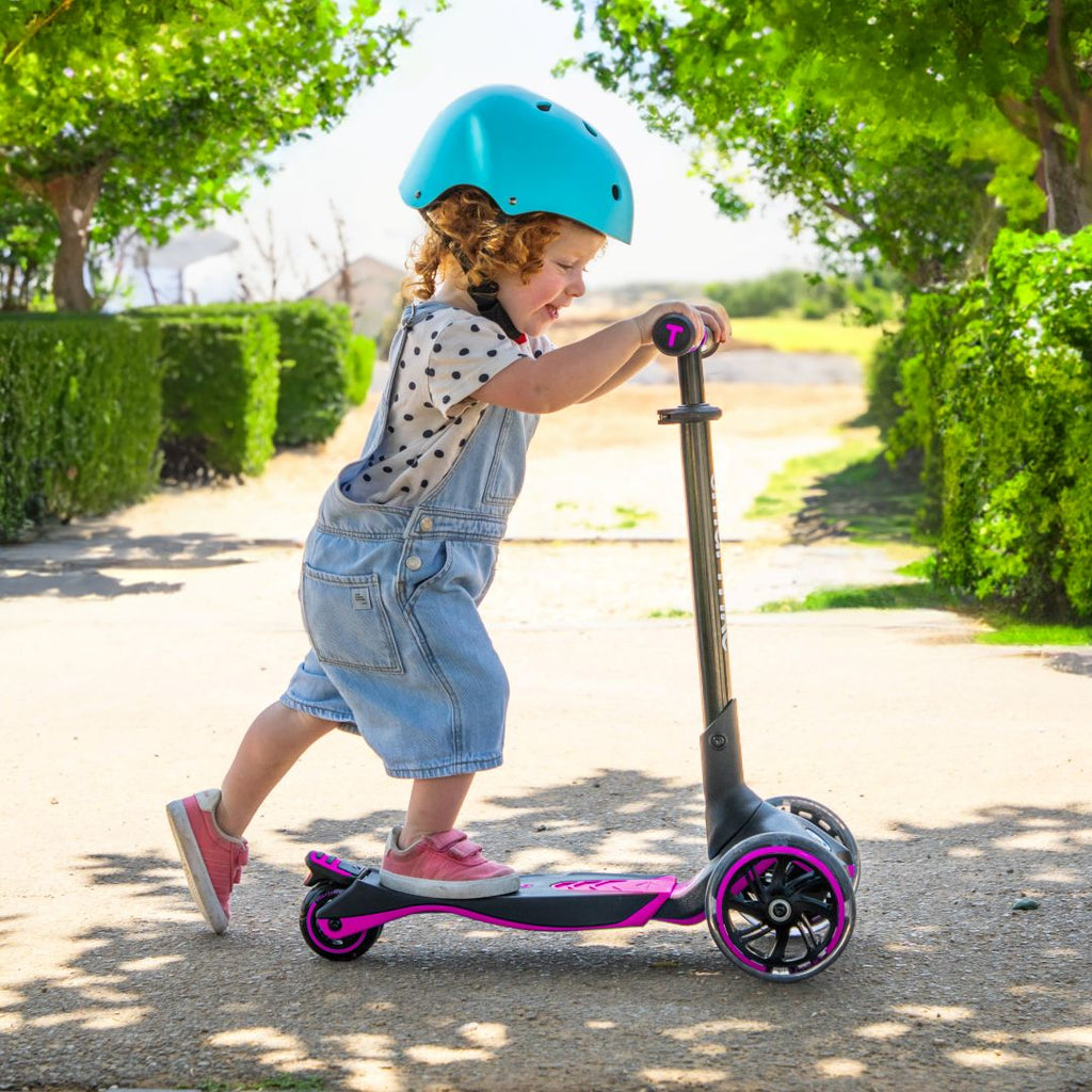 Bebé de aproximadamente 2 años con casco turquesa impulsándose con energía en el scooter evolutivo SmarTrike Xtend rosa en etapa inicial, en un sendero con vegetación y luz solar de fondo.