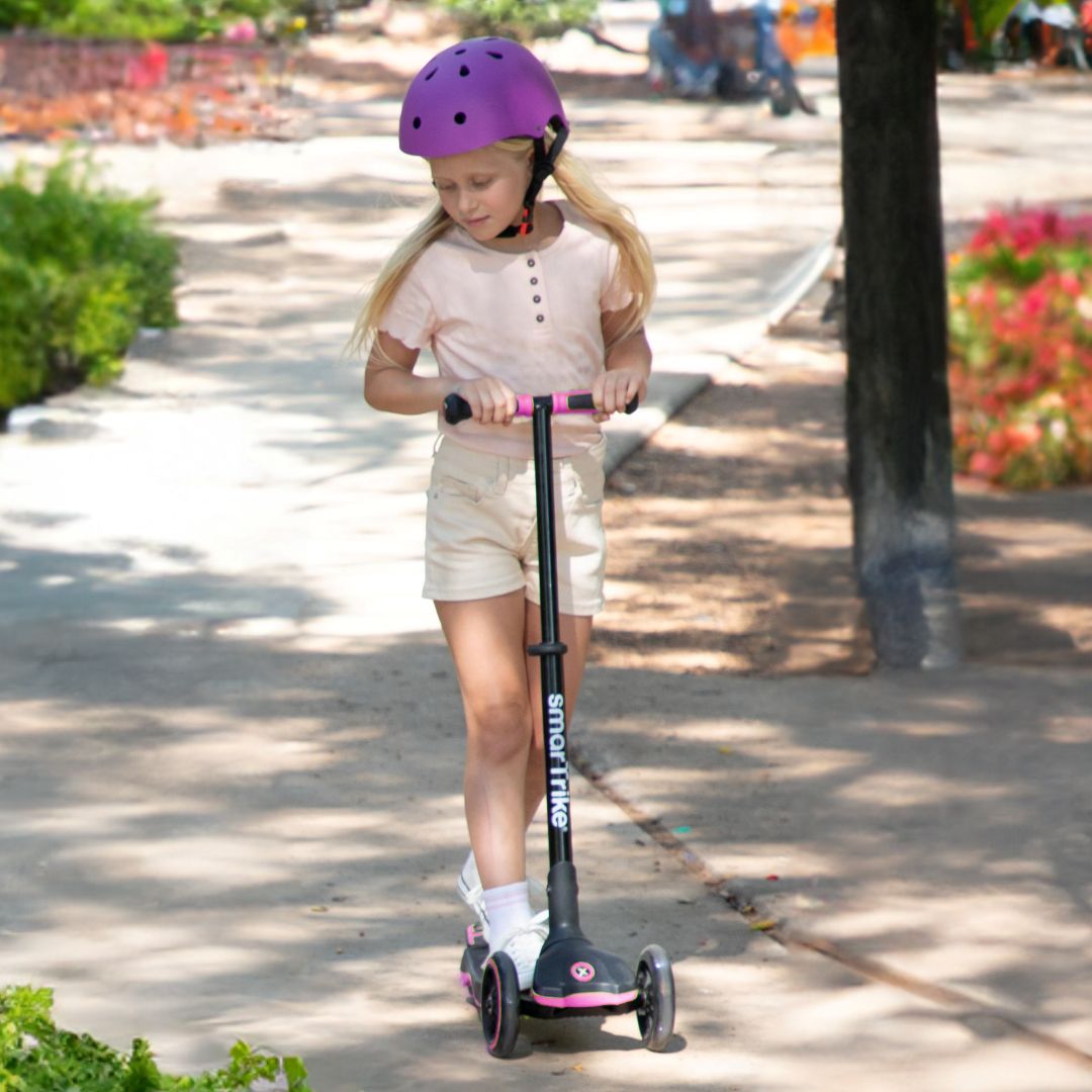 Niña de aproximadamente 7 años con casco morado manejando el scooter evolutivo SmarTrike Xtend en color rosa en modo scooter, en un sendero de parque soleado con vegetación de fondo.