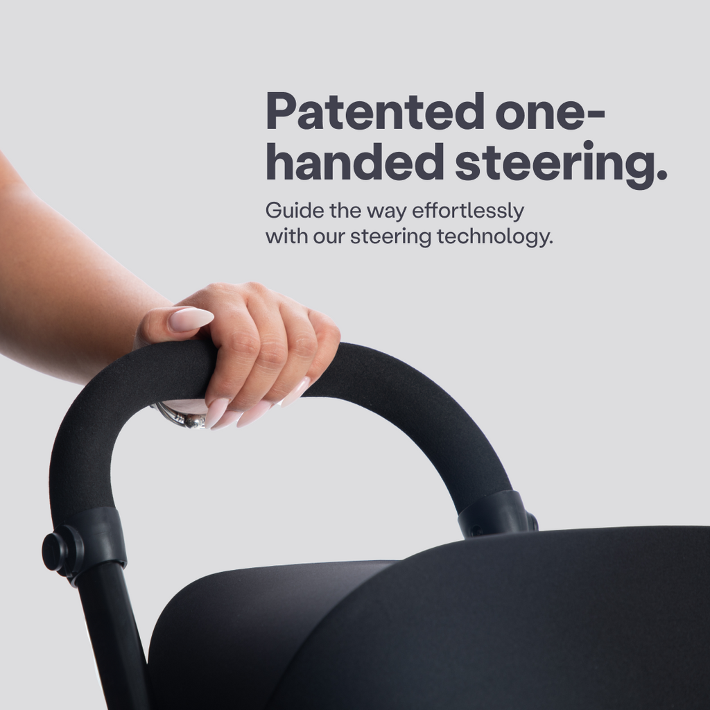 Detalle de una mano sujetando con una sola mano el manubrio ergonómico negro del coche triciclo SmarTrike Wonder, destacando el sistema de dirección con una mano patentado. Texto en la imagen: "Patented one-handed steering. Guide the way effortlessly with our steering technology."