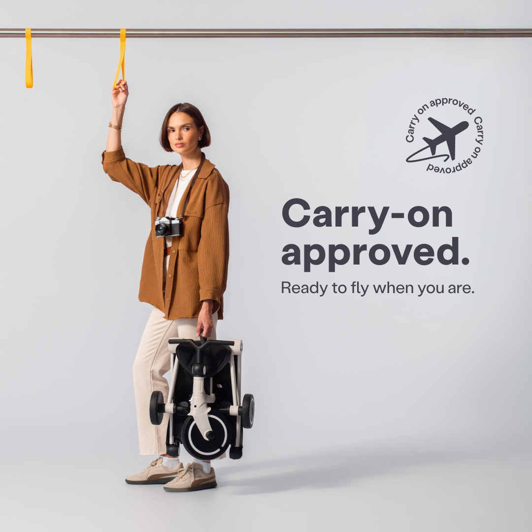 Mujer de pie sosteniendo con una mano el coche triciclo SmarTrike Wonder plegado en color blanco, y con la otra mano un pasamano amarillo de metro o tren. Lleva una cámara de fotos colgada al cuello. Texto en la imagen: "Carry-on approved. Ready to fly when you are." con ícono de avión. Wonder es apto para cabina de avión, plegado compacto.
