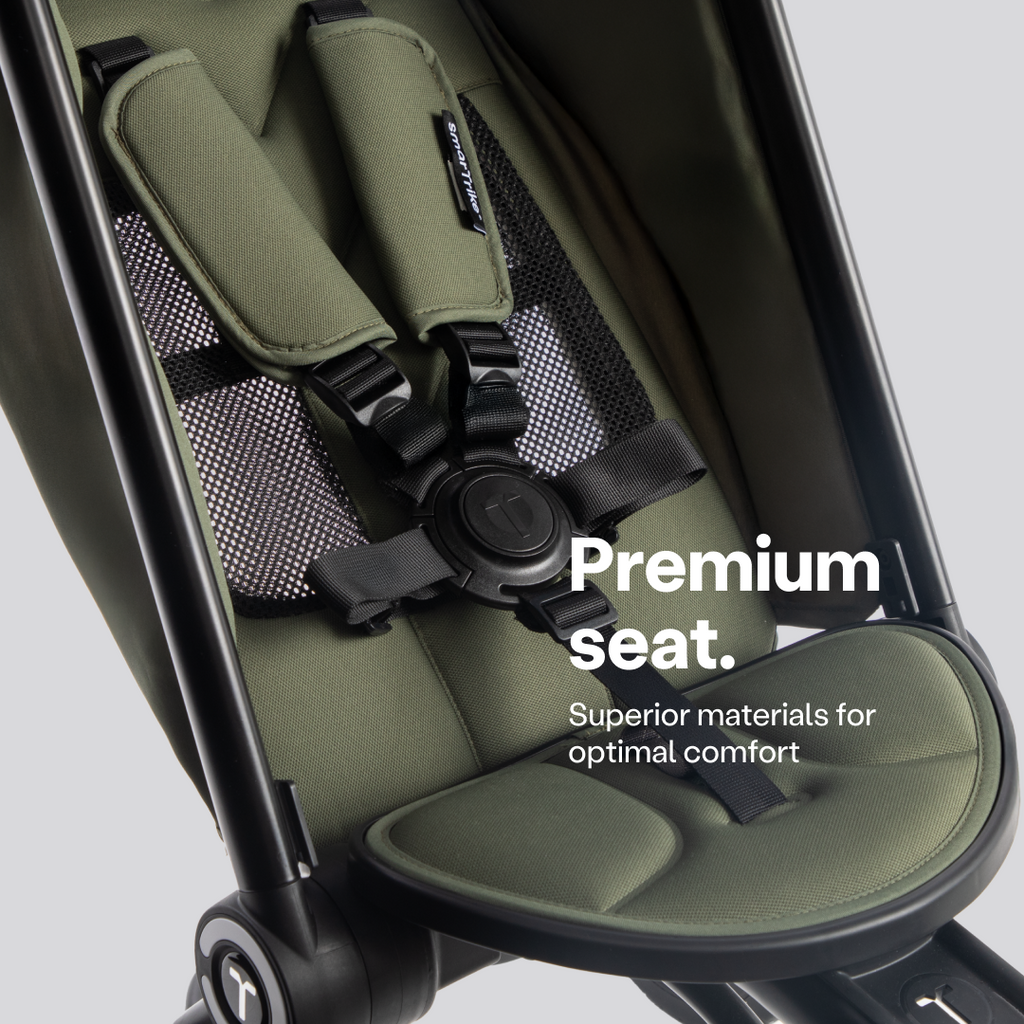 Detalle del asiento del coche triciclo SmarTrike Wonder max en color verde oliva, mostrando el arnés de seguridad de 5 puntos con hebilla central, acolchado suave, respaldo de malla transpirable y reposapiés ergonómico. Texto en la imagen: "Premium seat. Superior materials for optimal comfort."