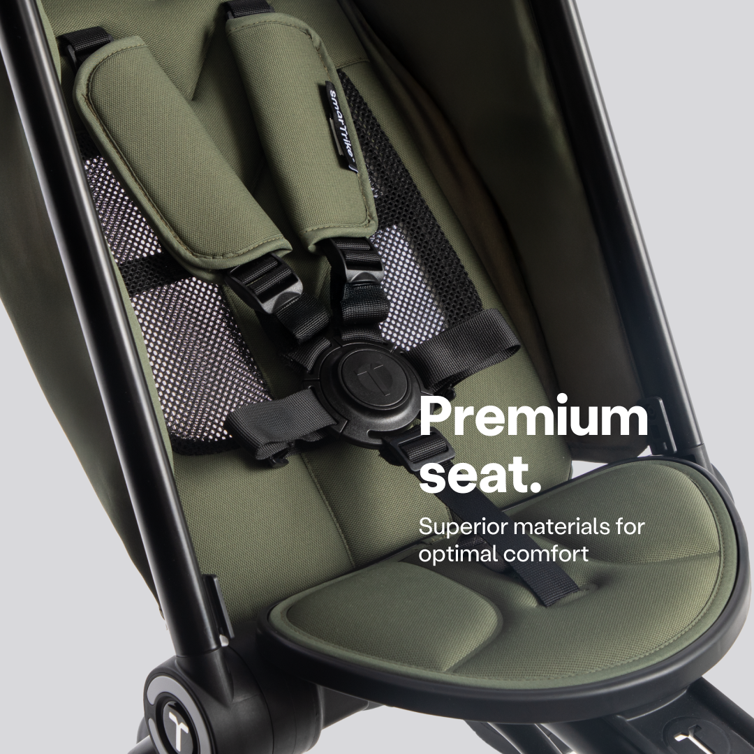 Detalle del asiento del coche triciclo SmarTrike Wonder max en color verde oliva, mostrando el arnés de seguridad de 5 puntos con hebilla central, acolchado suave, respaldo de malla transpirable y reposapiés ergonómico. Texto en la imagen: "Premium seat. Superior materials for optimal comfort."