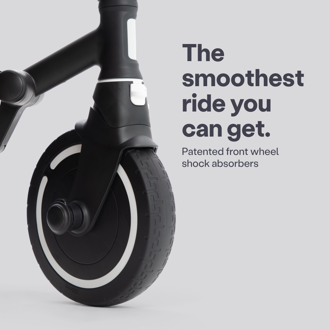 Detalle de la rueda delantera negra con aro blanco del coche triciclo SmarTrike Wonder max, destacando el sistema de amortiguación frontal patentado. Texto en la imagen: "The smoothest ride you can get. Patented front wheel shock absorbers."