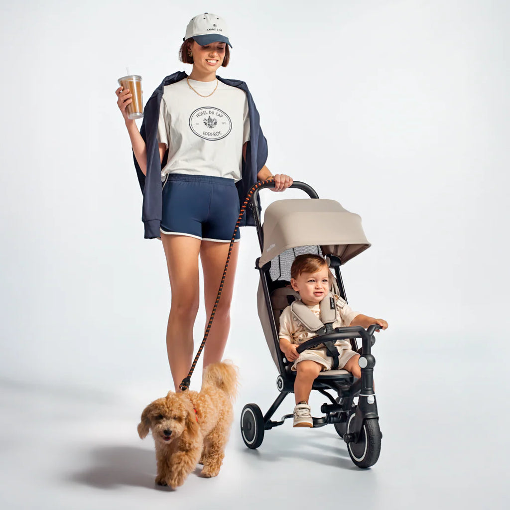 Mamá sonriente paseando con un café en mano y su perro con correa, mientras un bebé va sentado en el coche triciclo evolutivo SmarTrike Wonder max en color beige con estructura negra, con toldo solar y barra de seguridad delantera. Fondo gris claro.