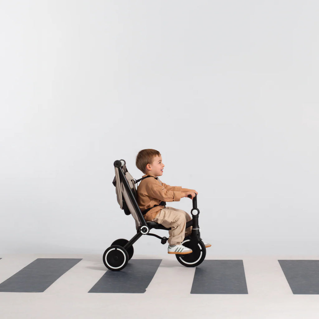 Niño sonriente de aproximadamente 3 años pedaleando de forma autónoma el coche triciclo evolutivo SmarTrike Wonder max en color beige con estructura negra, en modo triciclo sin toldo. Suelo con patrón de cuadros en gris y blanco, fondo gris claro.