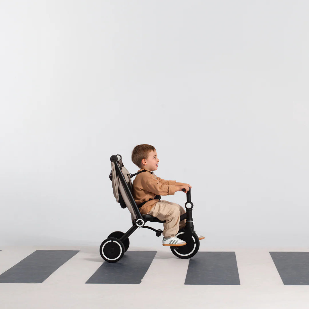 Niño sonriente de aproximadamente 3 años pedaleando de forma autónoma el coche triciclo evolutivo SmarTrike Wonder max en color beige con estructura negra, en modo triciclo sin toldo. Suelo con patrón de cuadros en gris y blanco, fondo gris claro.