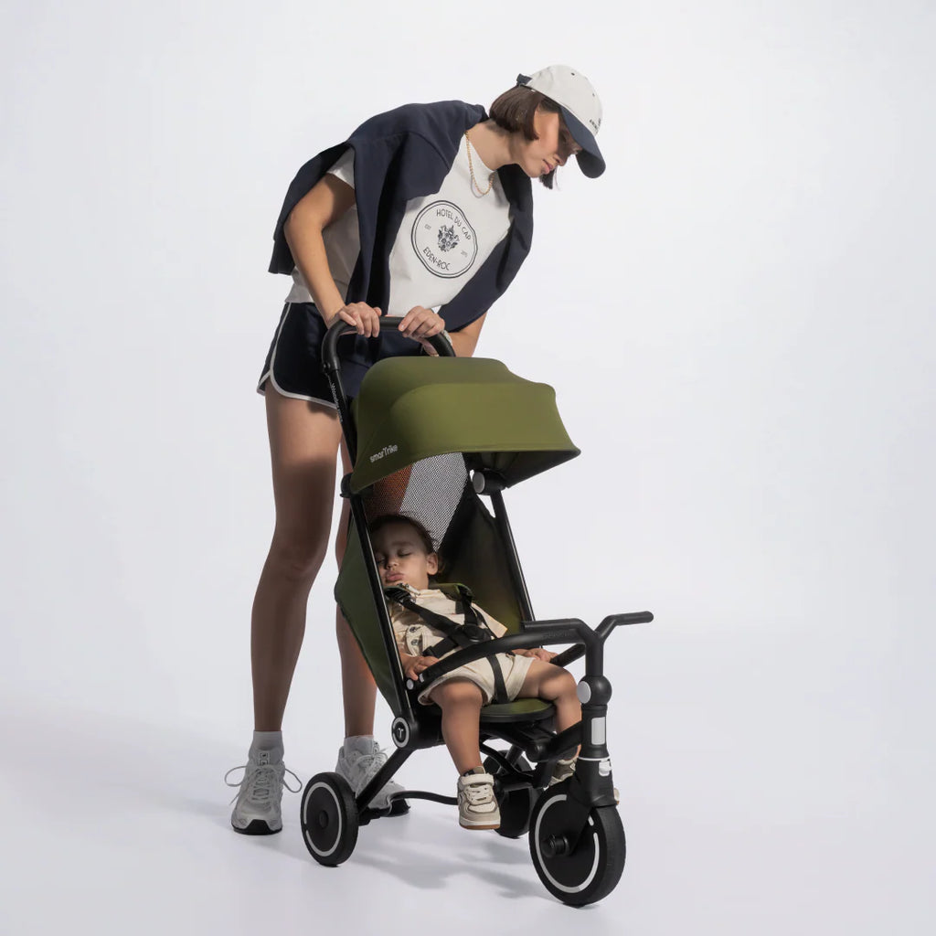 Mamá inclinándose a mirar a su bebé dormido en el coche triciclo evolutivo SmarTrike Wonder max en color verde oliva con estructura negra, con toldo solar extendido y arnés de seguridad. Fondo gris claro.