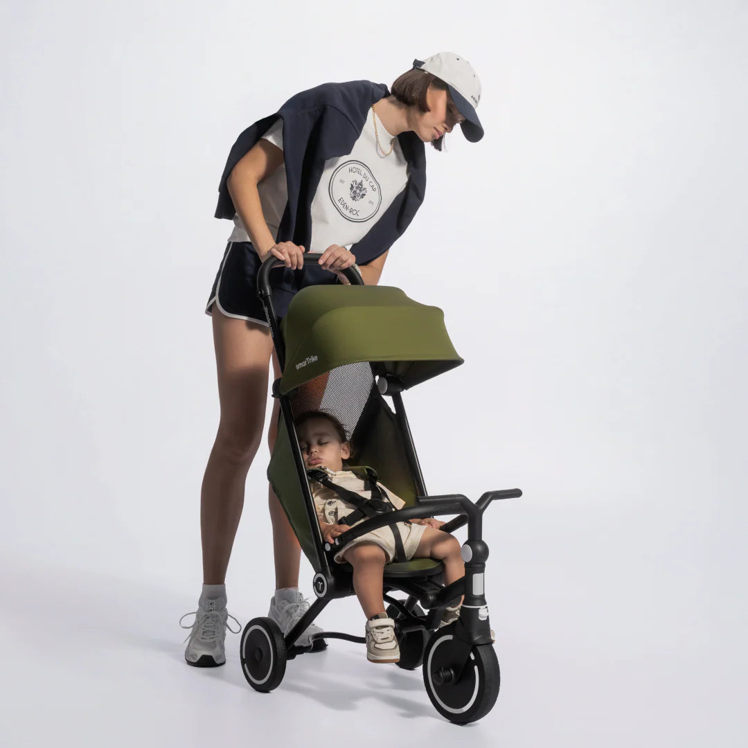 Mamá inclinándose a mirar a su bebé dormido en el coche triciclo evolutivo SmarTrike Wonder max en color verde oliva con estructura negra, con toldo solar extendido y arnés de seguridad. Fondo gris claro.