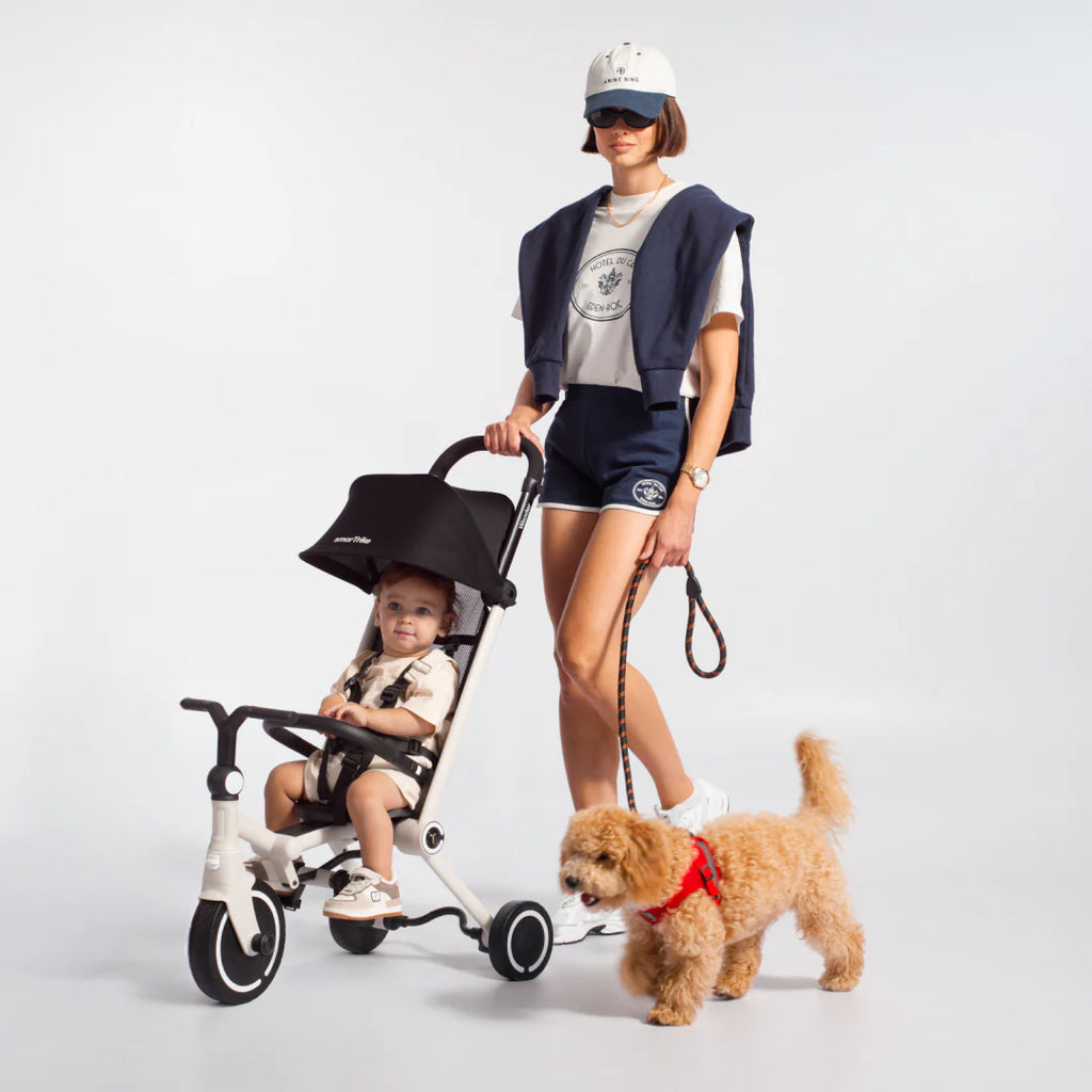 Mamá de pie paseando con su perro con correa, mientras un bebé va sentado con arnés en el coche triciclo evolutivo SmarTrike Wonder en color blanco con toldo negro desplegado y barra delantera. Fondo gris claro.