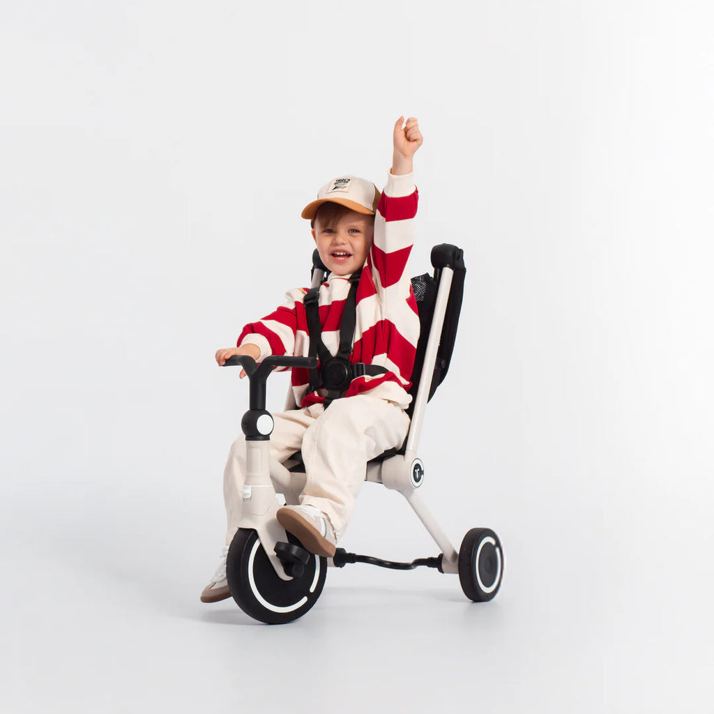 Niño feliz con el brazo levantado en señal de celebración, sentado en el coche triciclo evolutivo SmarTrike Wonder en color blanco con estructura negra, en modo triciclo sin toldo y con arnés de seguridad puesto. Fondo gris claro. 