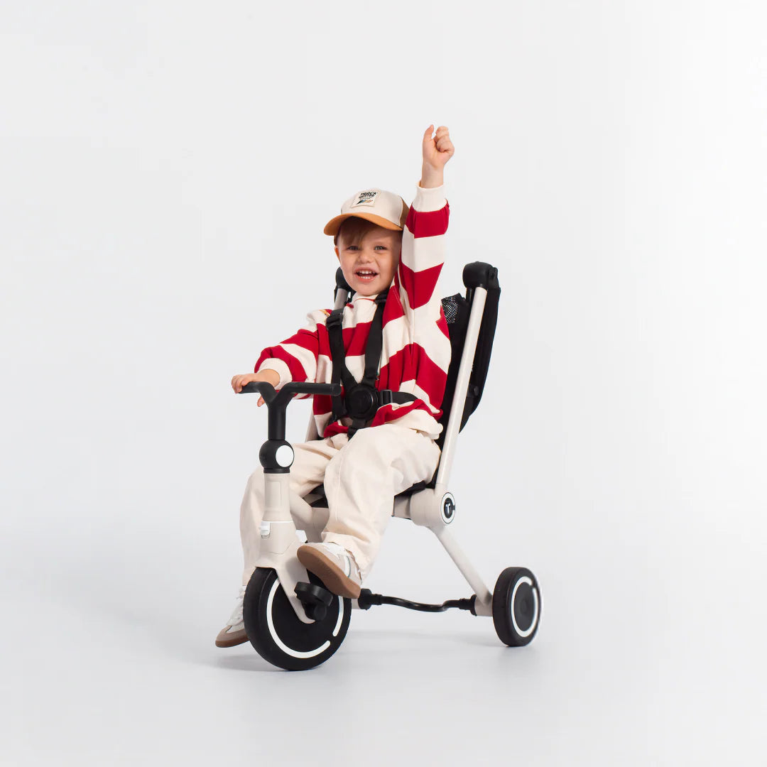 Niño feliz con el brazo levantado en señal de celebración, sentado en el coche triciclo evolutivo SmarTrike Wonder en color blanco con estructura negra, en modo triciclo sin toldo y con arnés de seguridad puesto. Fondo gris claro. 
